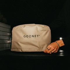 A beige canvas dust bag with the brand name 'GOZNEY' printed oven cover for Arc Lite Oven in black on the front, resting on a dark surface with a person's hand resting near it against a dark background.