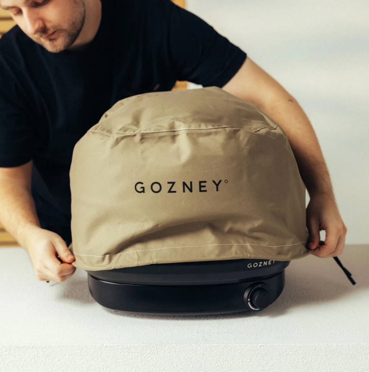 A man wearing a black t-shirt is shown from the side as he adjusts a beige protective oven cover on a black portable stove. The cover has the brand name 'GOZNEY' printed in black on the front.