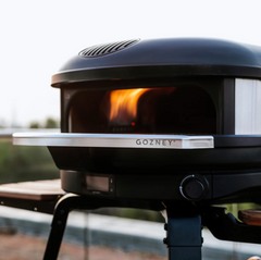 A close-up of a black GOZNEY outdoor pizza oven with a visible fire inside, set on a table outdoors with a blurred background.