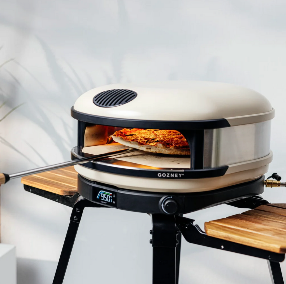 A Gozney portable outdoor pizza oven on a tripod stand, with a pizza baking inside and a metal peel holding a utensil extended from the left. The oven has a cream-colored exterior, a black base and trim, and a stainless steel interior.