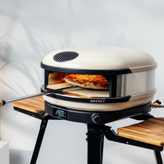 A Gozney portable outdoor pizza oven on a tripod stand, with a pizza baking inside and a metal peel holding a utensil extended from the left. The oven has a cream-colored exterior, a black base and trim, and a stainless steel interior.