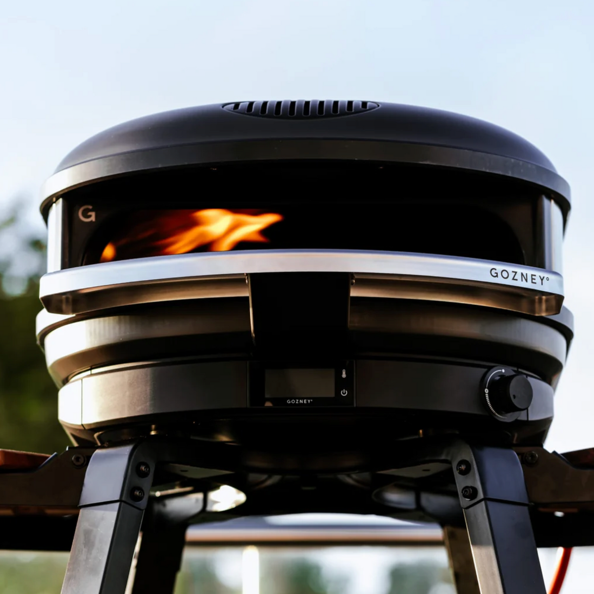 Close-up of a black GOZNEY outdoor fire pit grill with a visible flame inside, mounted on a metal stand against a blurred outdoor background.