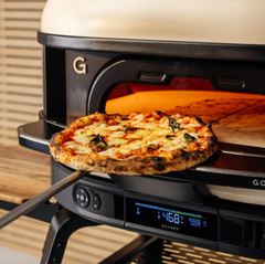 A close-up of a pizza being pulled out of a GOZNEY gas wood-fired pizza oven using a long metal peel. The oven has a cream-colored ceramic dome, a black metal frame, and a digital control panel on the front with a glowing orange interior.