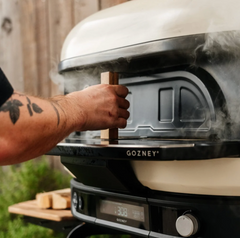 A person's arm is shown pushing a wooden release lever on a black and cream Gozney Smitter grill, with smoke rising from the cooking chamber in an outdoor setting.