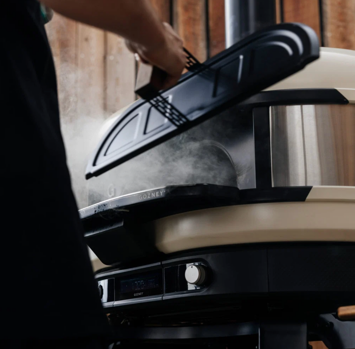 A close-up lifestyle shot of a person lifting the lid of a black and beige outdoor kamado grill, with steam rising from inside. The background shows a rustic wooden wall.