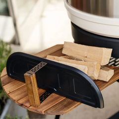 A close-up of a black metal fire starter guard resting on a round wooden slatted tray, with several light wood firewood logs placed on top. A white and black cooking vessel is partially visible in the upper right, and the background is softly blurred.