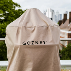 A beige outdoor grill cover with the brand name 'GOZNEY°' printed in black across the center, shown outdoors with a blurred city skyline and green trees in the background.