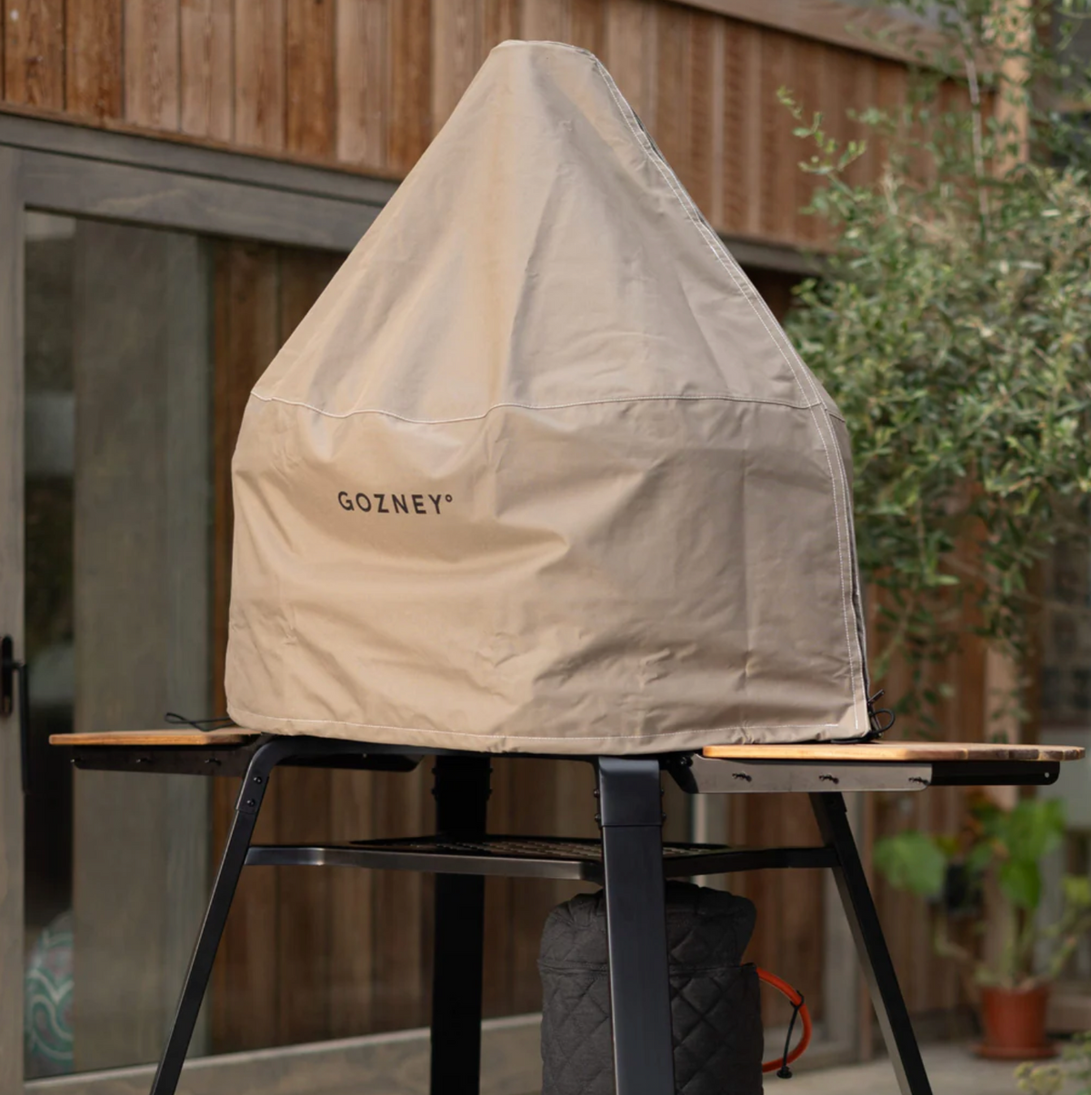 A beige outdoor grill cover with the brand name 'GOZNEY' printed on the front, draped over a tabletop grill in a patio setting.