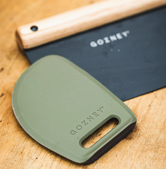 An olive green GOZNEY silicone scraper and a black scraper blade with a wooden handle, both branded GOZNEY, displayed on a wooden surface.