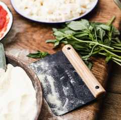 A dark gray flour scraper with a light wood handle, branded GOZNEY, resting on a rustic wooden cutting board surrounded by baking ingredients like flour, fresh basil, and cheese.