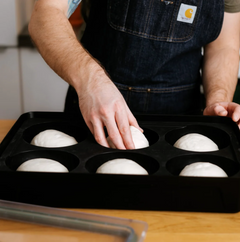 A person wearing a dark denim apron places a round white dough ball into a black six-cup baking tray on a wooden table.