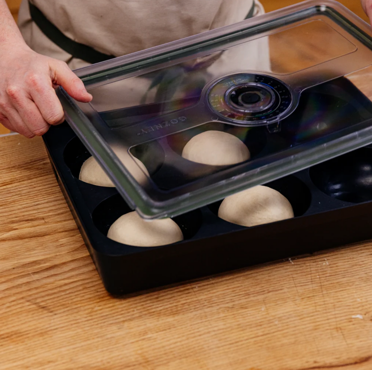 A hand lifts a clear plastic lid from a black oval baking tray containing four pale dough balls, set on a wooden surface.