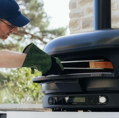 A person wearing a navy blue cap and a green oven mitt inserts a pizza stone into a black GOZNEY outdoor pizza oven. The scene is set outdoors with trees and a stone wall in the background.