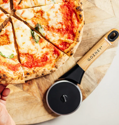 A pizza cutter with a black circular blade and a light wood handle engraved with 'GOZNEY' is shown beside a sliced pizza on a round wooden serving board. A hand is visible at the lower left edge.