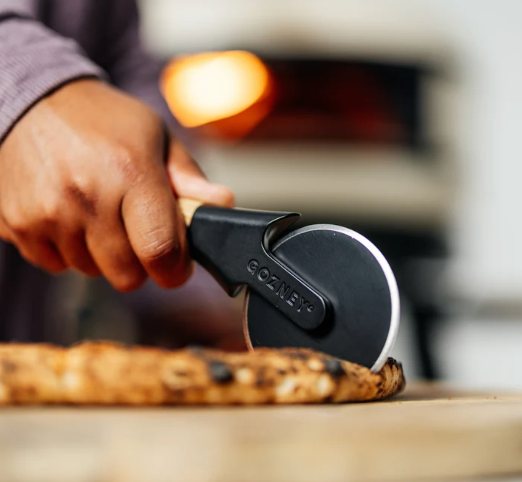 A close-up of a hand using a GOZANE pizza cutter to slice a freshly baked pizza on a wooden surface, with a blurred pizza oven in the background.