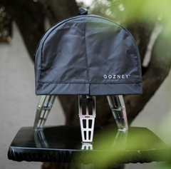 A black outdoor grill cover with the GOZNEY brand name printed in white on the front, displayed on a black table with a blurred tree background.