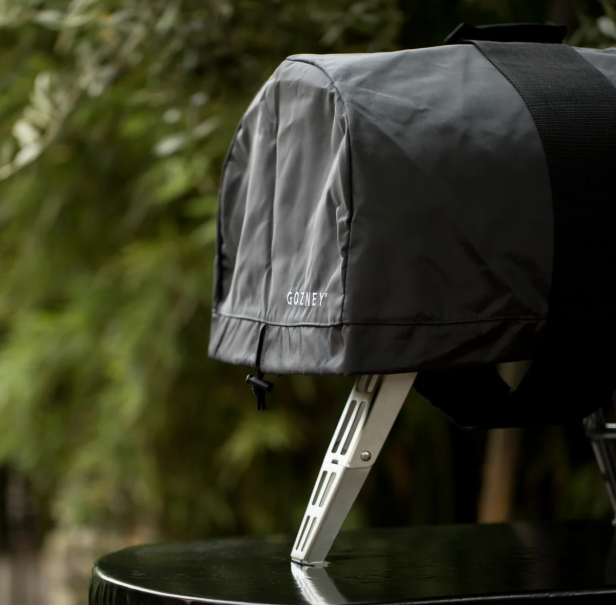 A dark gray protective grill cover with the brand name 'COOZY' printed in white, draped over a grill on a black table with a blurred green outdoor background.