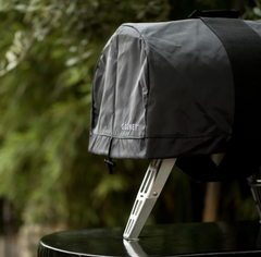 A dark gray protective grill cover with the brand name 'COOZY' printed in white, draped over a grill on a black table with a blurred green outdoor background.