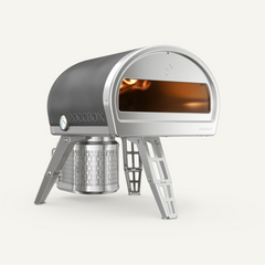 A Roccbox Soznet wood-burning pizza oven with a dark gray dome-shaped exterior, silver metal legs, and a visible fire inside the oven window, set against a solid off-white background.