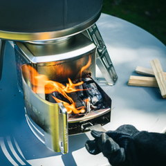 A close-up outdoor shot of a metallic portable stove with a fire burning inside, set on a round table. A gloved hand is adjusting the front panel, and small wood pieces sit to the right.