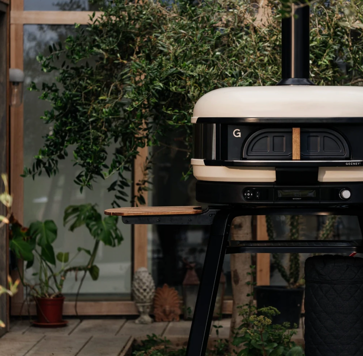 A modern outdoor pizza oven on a black metal stand, set on a patio with a wooden side table and lush green plants in the background.