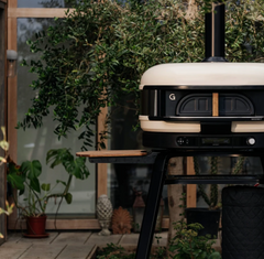 A modern outdoor pizza oven on a black metal stand, set on a patio with a wooden side table and lush green plants in the background.