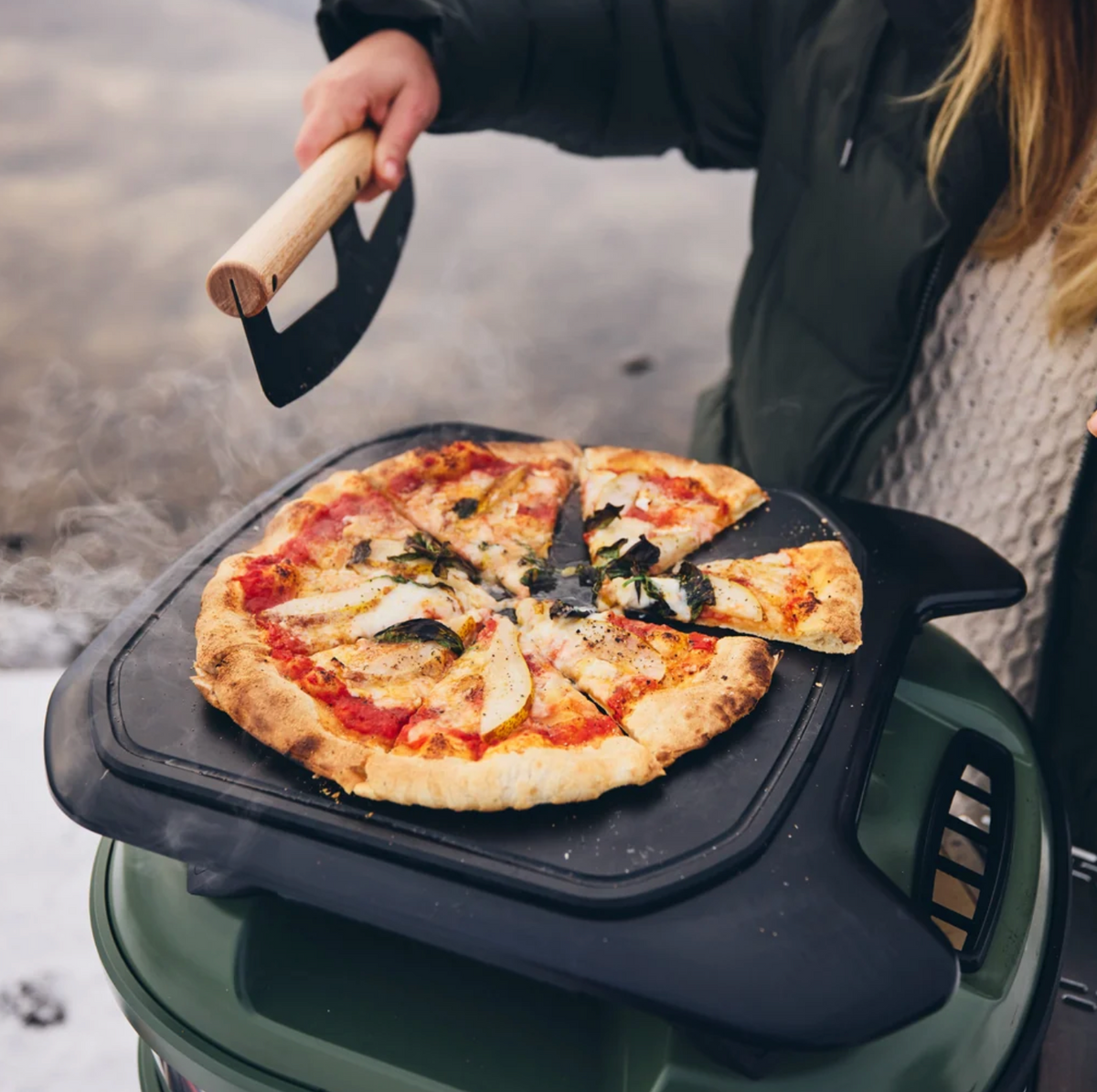 A person wearing a dark jacket is cooking a pizza on a black outdoor griddle attachment mounted on a green portable stove, with steam rising in a real outdoor setting.