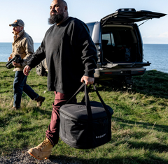 A man outdoors by the ocean carries a black Gozert storage bag in one hand while walking beside another person and an open van. The Gozert logo is visible on the bag.