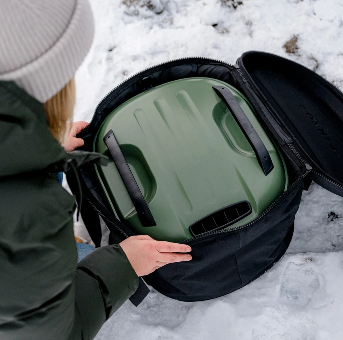 A person in a dark green puffer jacket and light gray beanie opens a black zippered bag in the snow, revealing an olive green insulated container with black handles inside.