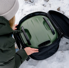A person in a dark green puffer jacket and light gray beanie opens a black zippered bag in the snow, revealing an olive green insulated container with black handles inside.