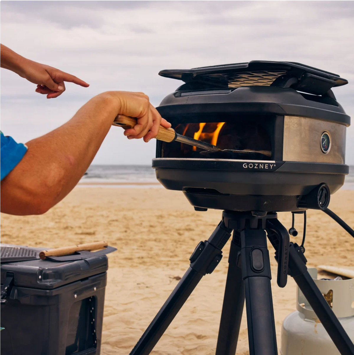 A Gozney outdoor electric smoker on a tripod set on a sandy beach, with a person's hands using a long handle to turn the fire inside the smoker. A cooler and propane tank are visible in the background.
