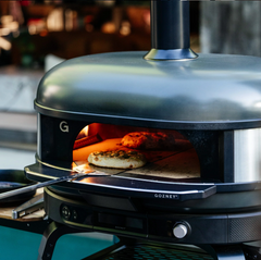 A Gozney outdoor pizza oven with its door open, showing a pizza baking inside. The oven has a matte black finish with a brushed silver dome lid and a visible brand logo 'G' on the side.