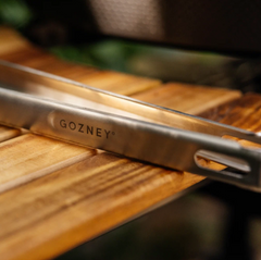Close-up of stainless steel tongs engraved with the brand name GOZNEY, resting on a wooden surface.
